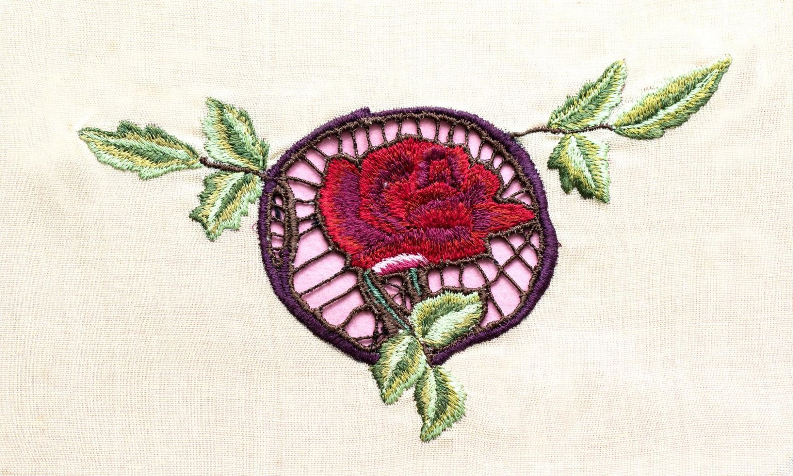 An embroidered design on fabric features a red rose at the center, surrounded by green leaves. The rose is enclosed within a circular frame with a lattice-like pattern. The background fabric is light beige, complementing the detailed stitching and vibrant colors of the embroidery.