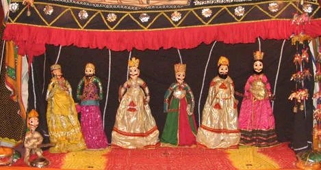 Six traditional puppets dressed in elaborate costumes are arranged in a row on a decorated stage. The backdrop features a red curtain and a black background adorned with gold and silver embellishments. The puppets wear crowns and ornate robes, suggesting royal or significant characters. Hanging ornaments decorate the right side of the stage, emphasizing the intricate craftsmanship and cultural art form of traditional puppetry.