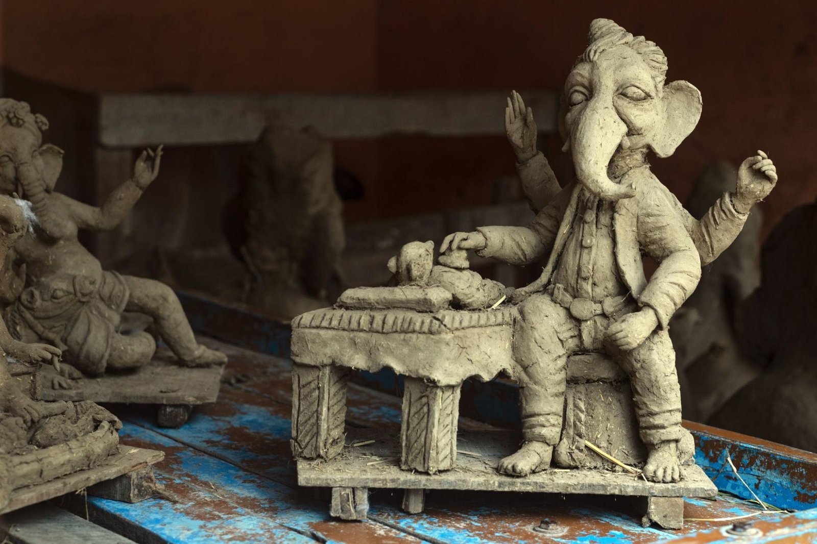 A collection of clay sculptures features the Hindu deity Ganesha, crafted with intricate details. The central figure is a seated Ganesha with multiple arms, adorned in traditional attire and holding symbolic objects. A table in front of the sculpture displays additional clay elements. Other Ganesha figures in different poses appear in the background, set against a blue wooden surface. The setting suggests an artisan's workshop, highlighting the craftsmanship and cultural significance of these sculptures.