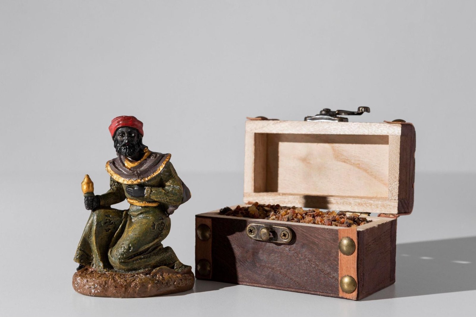 A small figurine of a person kneeling holds an object in one hand, positioned beside an open wooden box filled with amber-like pieces. The figurine is adorned in a green robe with gold accents and a red head covering. The wooden box has metal hinges and a latch, and its contents suggest historical or religious significance—possibly referencing one of the Three Wise Men from the Nativity story.