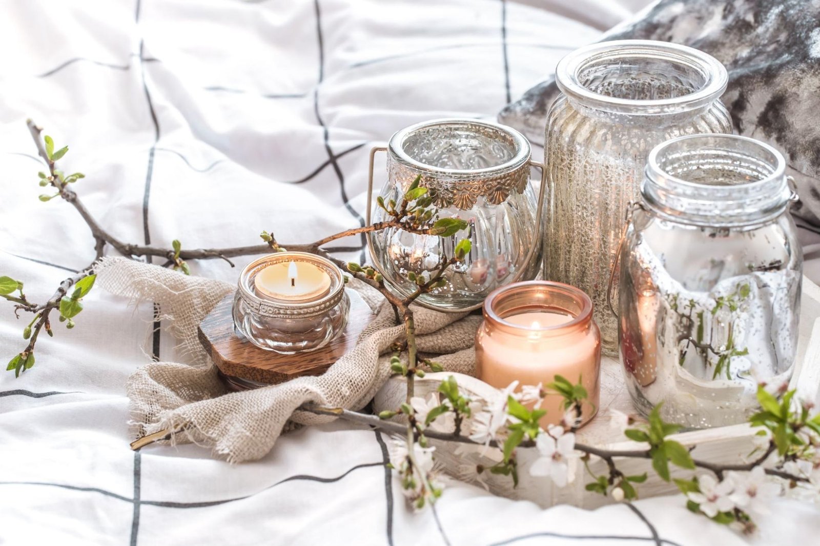A cozy arrangement of candles and glass jars is set on a bed with a white and black grid-patterned blanket. The display includes five candles in various holders—glass, copper, and metallic finishes—creating a warm ambiance. Branches with green leaves and white flowers surround the candles, adding a natural decorative touch. The setup exudes relaxation and aesthetic appeal, ideal for a serene atmosphere.