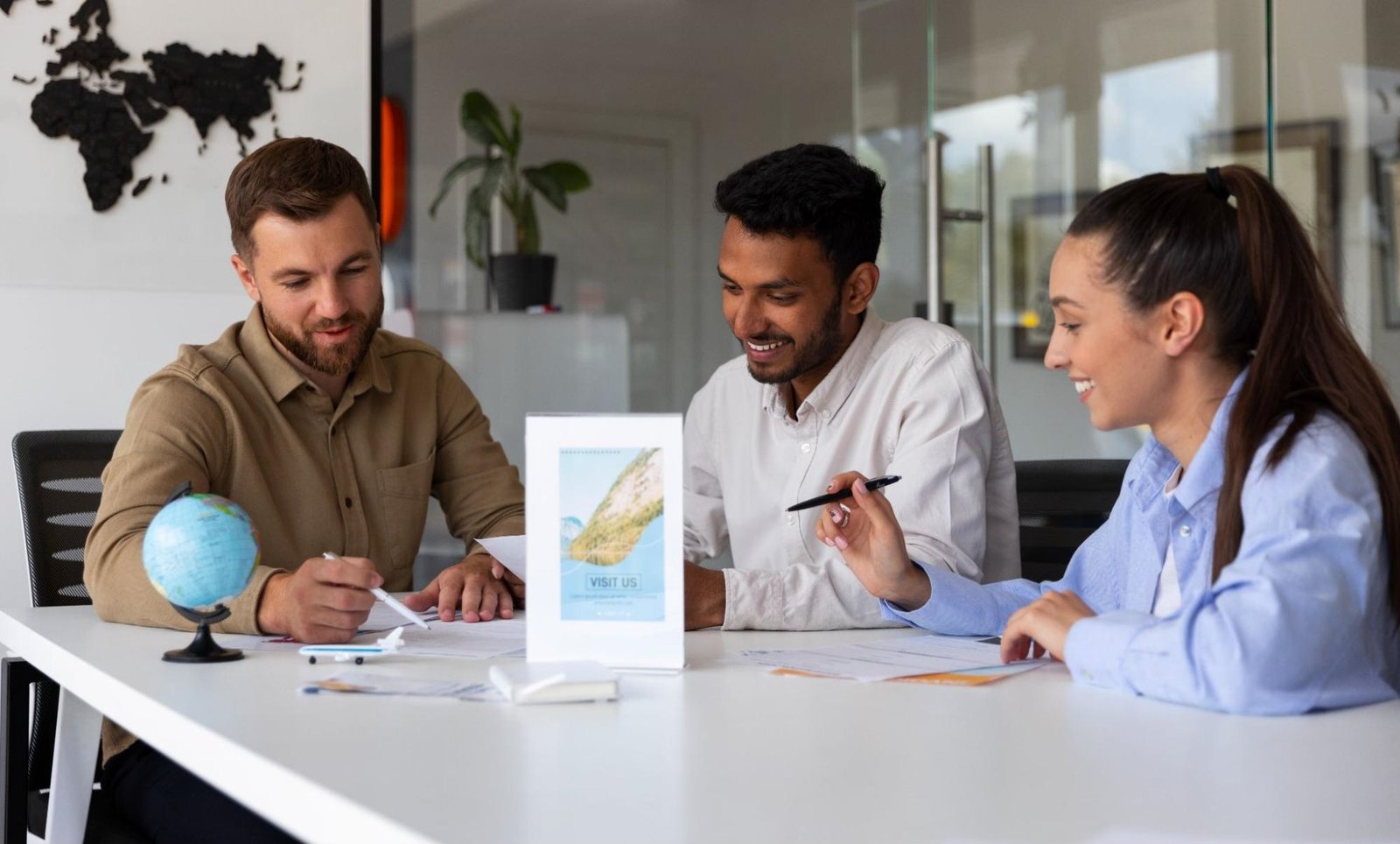 Three individuals sit at a table in an office or meeting room, engaged in a discussion. The table holds various items, including a small globe, papers, pens, and a stand displaying a promotional flyer labeled "VISIT US." The background features a world map on the wall and a potted plant, suggesting themes of travel, geography, or international collaboration.
