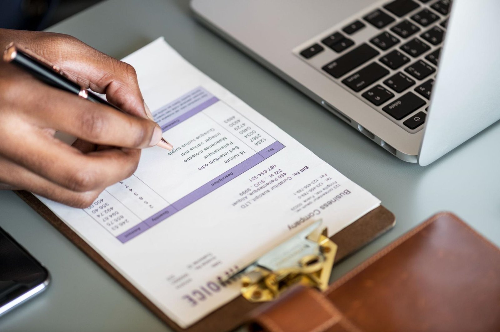 A person's hand holds a pen and writes on an invoice document placed on a clipboard. The invoice contains various details, including item descriptions, quantities, and prices, with a company name and customer information visible at the top. A laptop keyboard is in the background, along with a leather wallet or planner, emphasizing a business or financial setting.