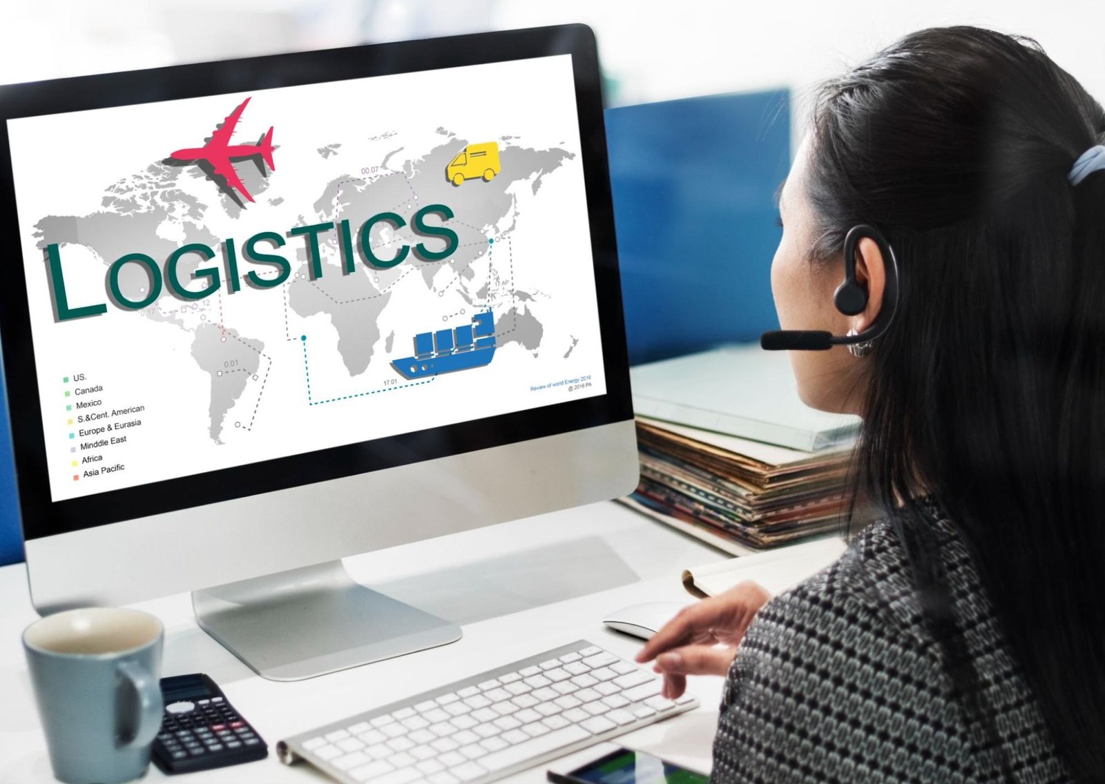 A person wearing a headset sits at a desk, looking at a computer screen displaying a world map with "LOGISTICS" written in large green letters. The map includes icons representing different transportation modes: an airplane, a delivery truck, and a cargo ship. A legend on the left side lists various regions, such as US, Canada, Mexico, S. & Central America, Europe & Eurasia, Middle East, Africa, and Asia Pacific. The scene suggests a logistics or supply chain professional coordinating transportation routes and methods in a global network.