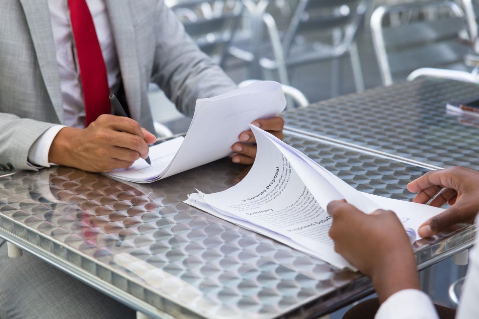Two individuals sitting at a metal table engaged in a business meeting. One person in a gray suit and red tie is signing documents, while the other hands over a proposal titled "IT Services Proposal." The setting appears to be an outdoor or semi-outdoor business environment, indicating a professional agreement or negotiation taking place.