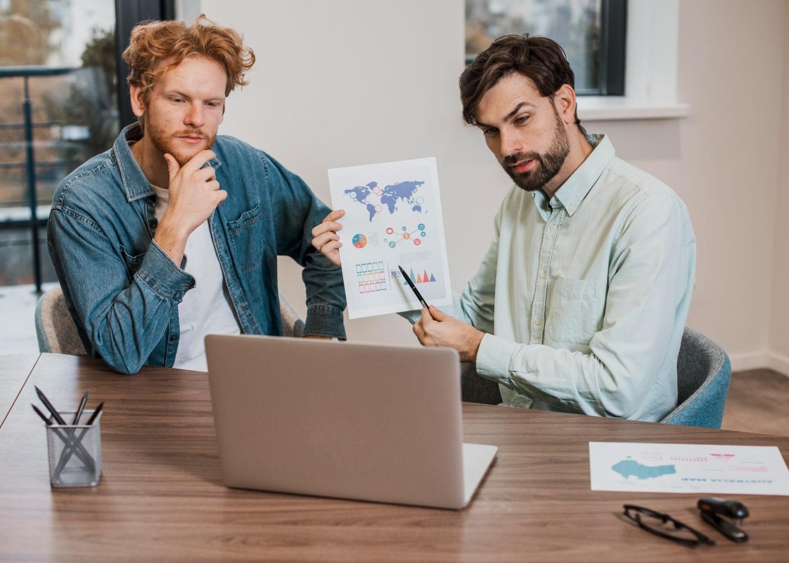 Two men are seated at a table in a modern office space, engaged in a discussion. One man in a light green shirt is holding and pointing to a printed sheet with colorful charts and a world map, while the other man in a denim shirt thoughtfully looks on. A laptop is open in front of them, suggesting they are analyzing global business or data trends during a meeting or virtual presentation.