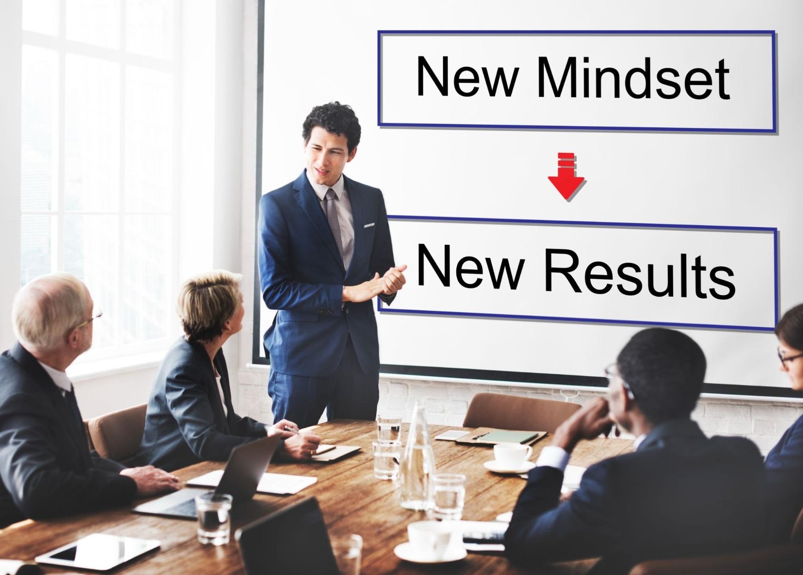 A business professional in a navy blue suit stands at the front of a meeting room, presenting to colleagues seated around a conference table. Behind him is a large screen displaying the motivational message "New Mindset → New Results" with a red downward arrow between the phrases. The setting suggests a corporate training session or strategic planning meeting.