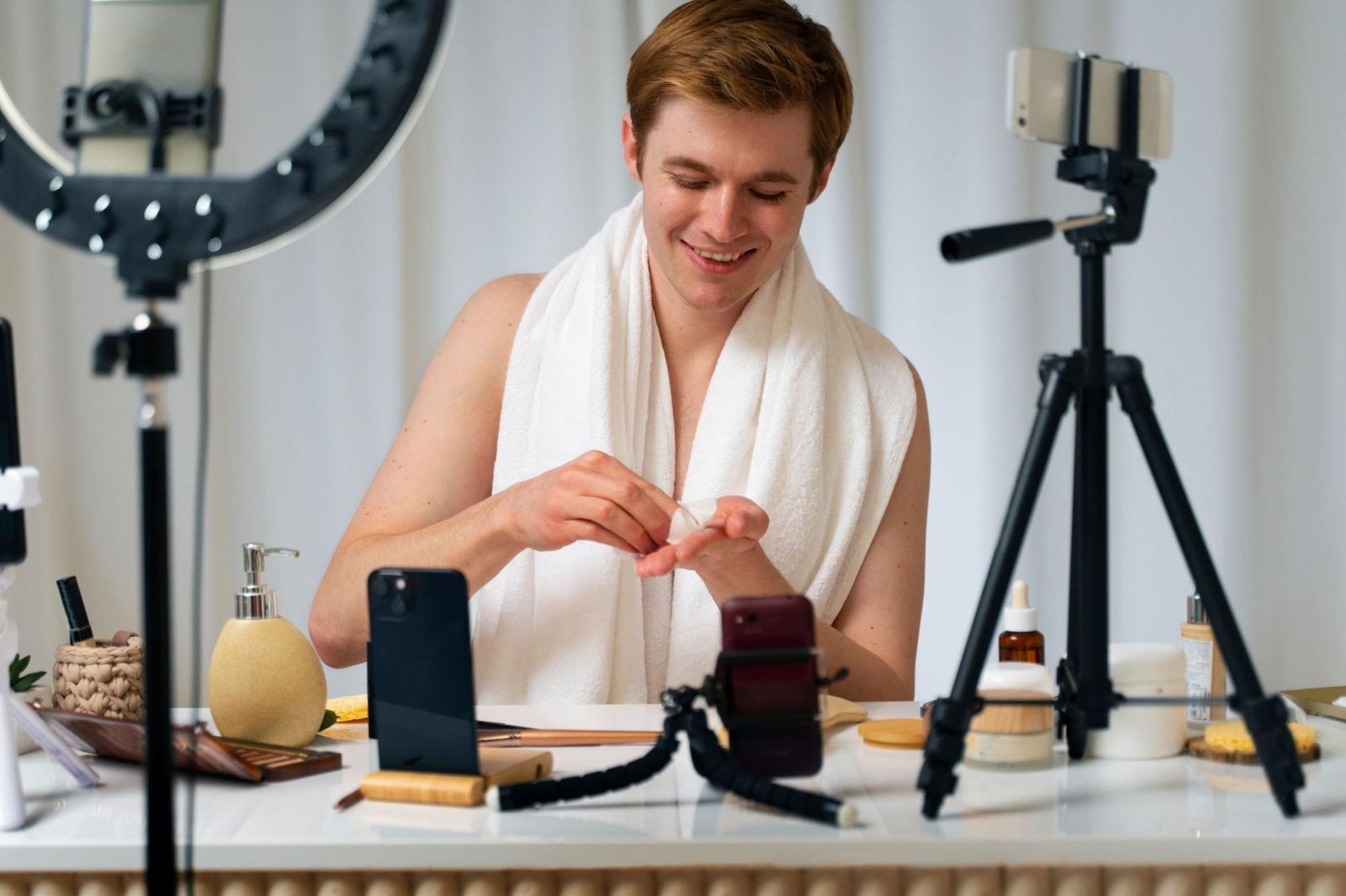 A young man with a white towel draped over his shoulders smiles while applying skincare product to his hand, seated at a table with various beauty and skincare items. Multiple smartphones mounted on tripods and a ring light surround him, indicating he is filming a skincare or grooming tutorial for social media or content creation.
