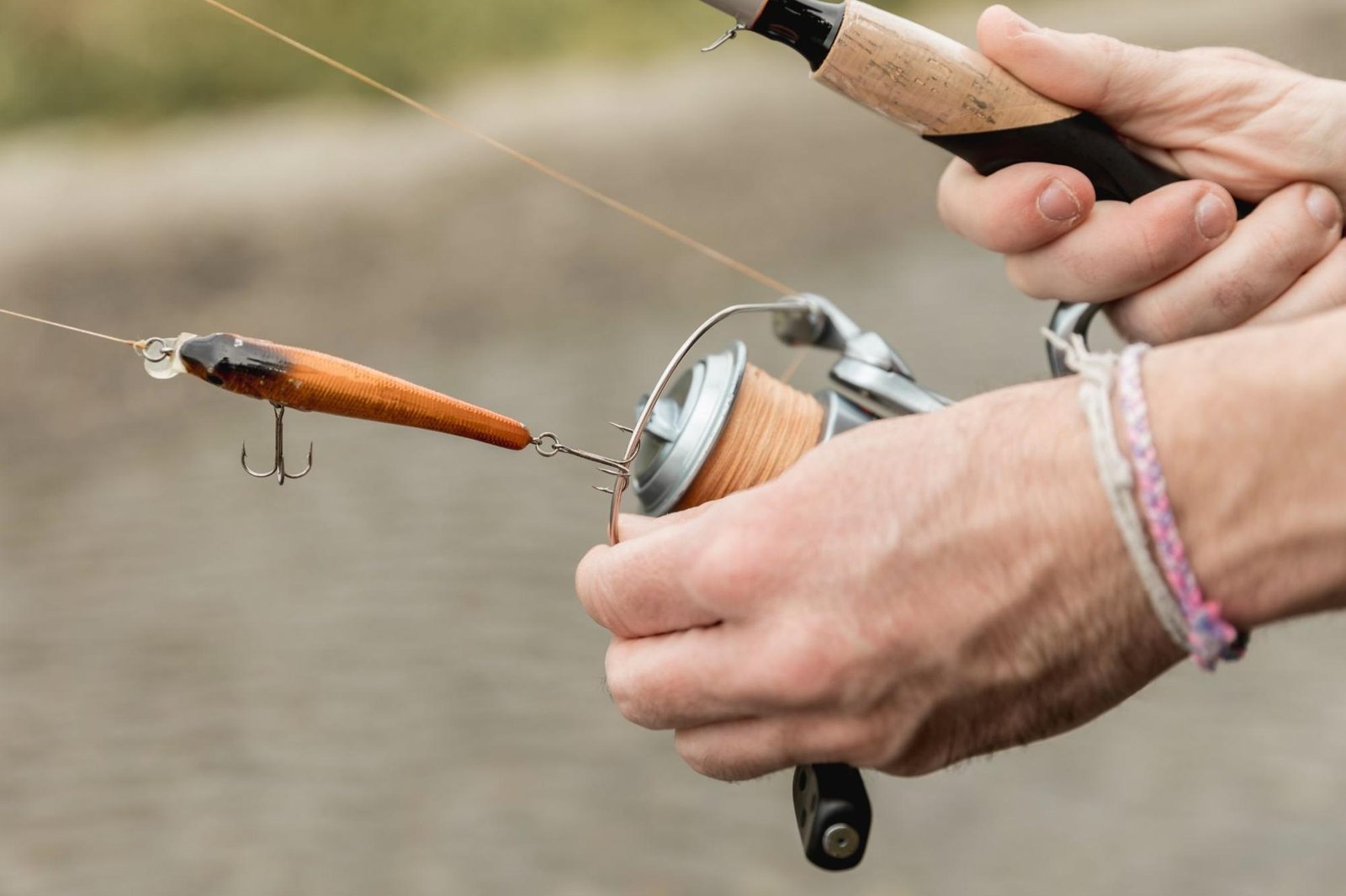 A close-up of a person preparing a fishing rod with an orange lure attached to the line, showcasing the reel, handle, and hand wearing a braided bracelet, with a blurred water background.
