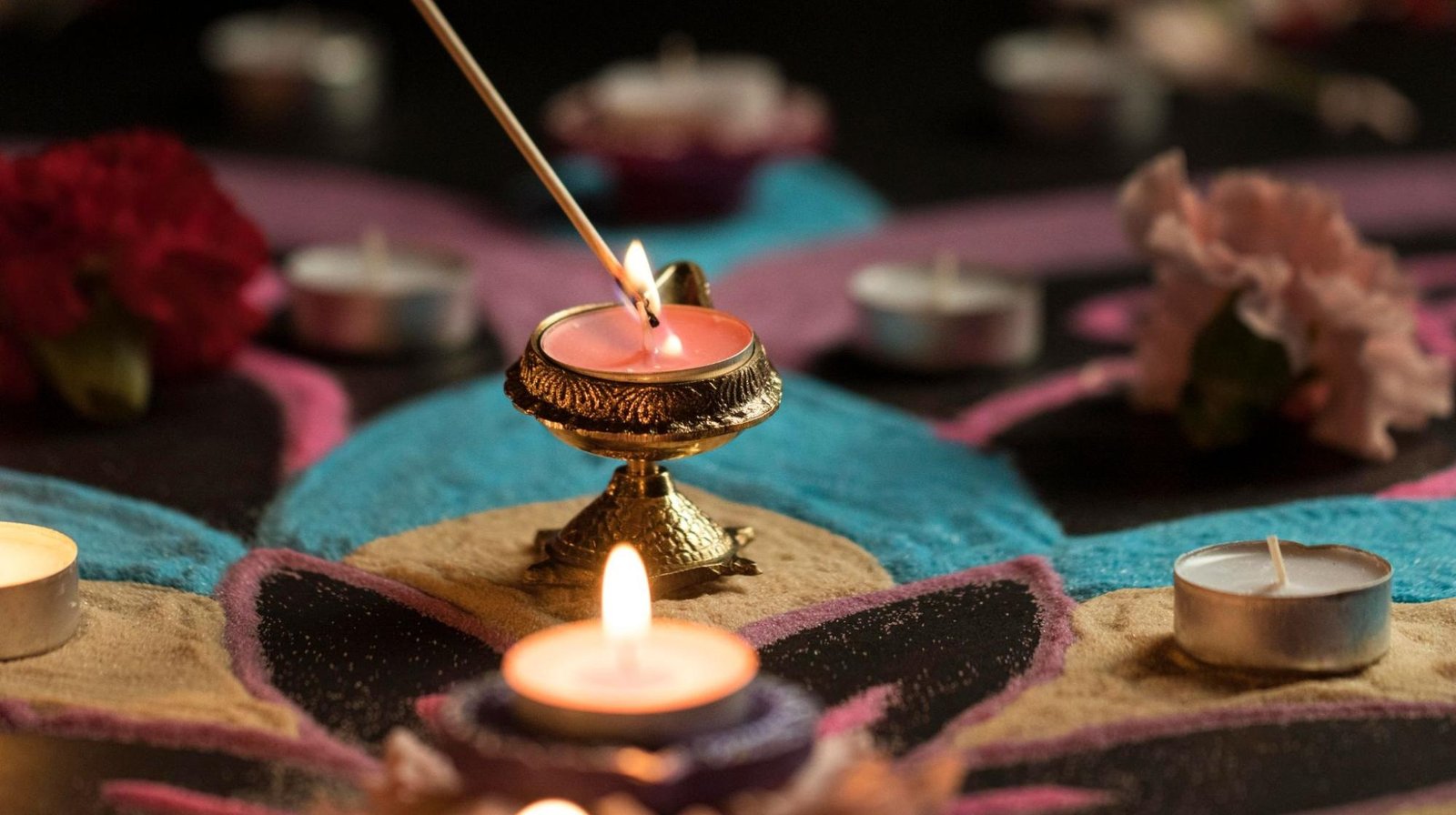 Close-up of a decorative Diya being lit with a matchstick, surrounded by tealight candles, colorful rangoli patterns, and flower petals, creating a festive and traditional ambiance.