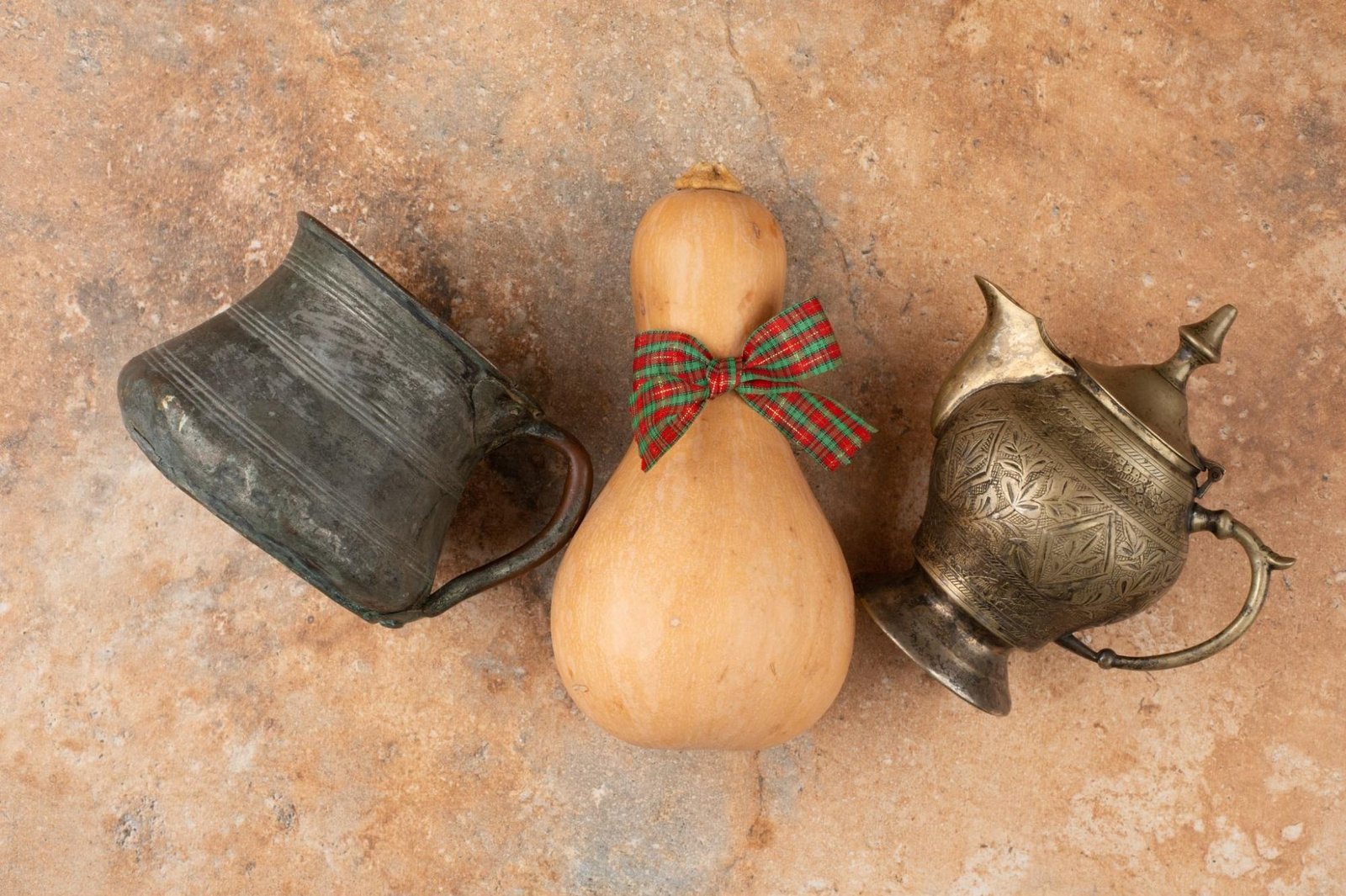 Top-down view of two antique metal vessels—one mug-like and one teapot-style—with a dried gourd placed between them, adorned with a red and green tartan ribbon bow, all set against a rustic stone background.