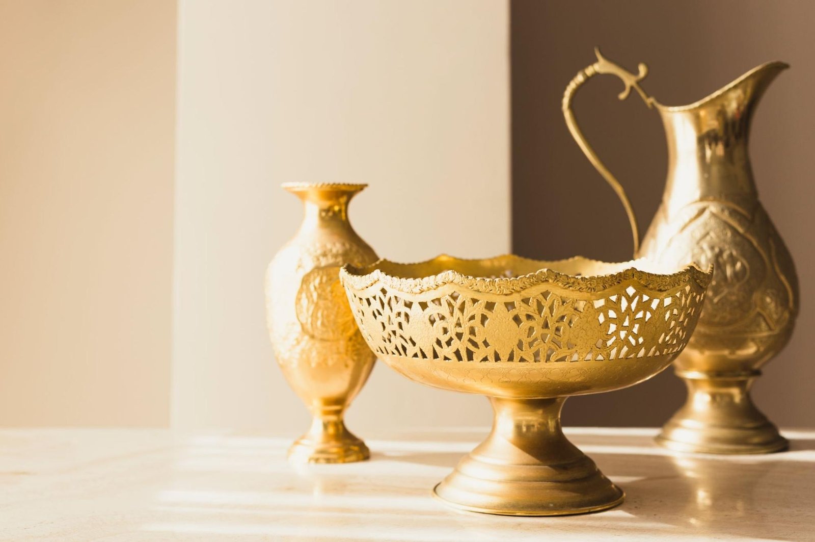 Three elegant golden vessels displayed on a marble surface under warm sunlight—an intricately carved bowl on a pedestal in the foreground, with a tall ornamental vase and a long-spouted pitcher featuring engraved patterns in the background.