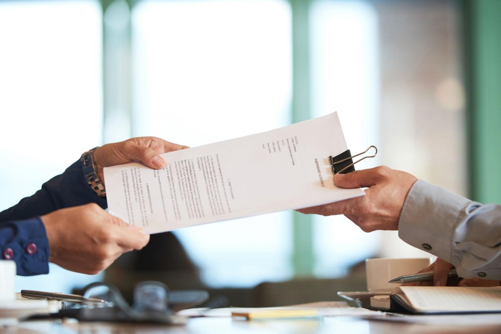 Close-up of two people exchanging a stack of printed documents secured with a binder clip across a desk. One person is handing over the papers while the other is receiving them. Office supplies, a notebook, and a coffee cup are visible on the table, suggesting a formal business or legal setting.