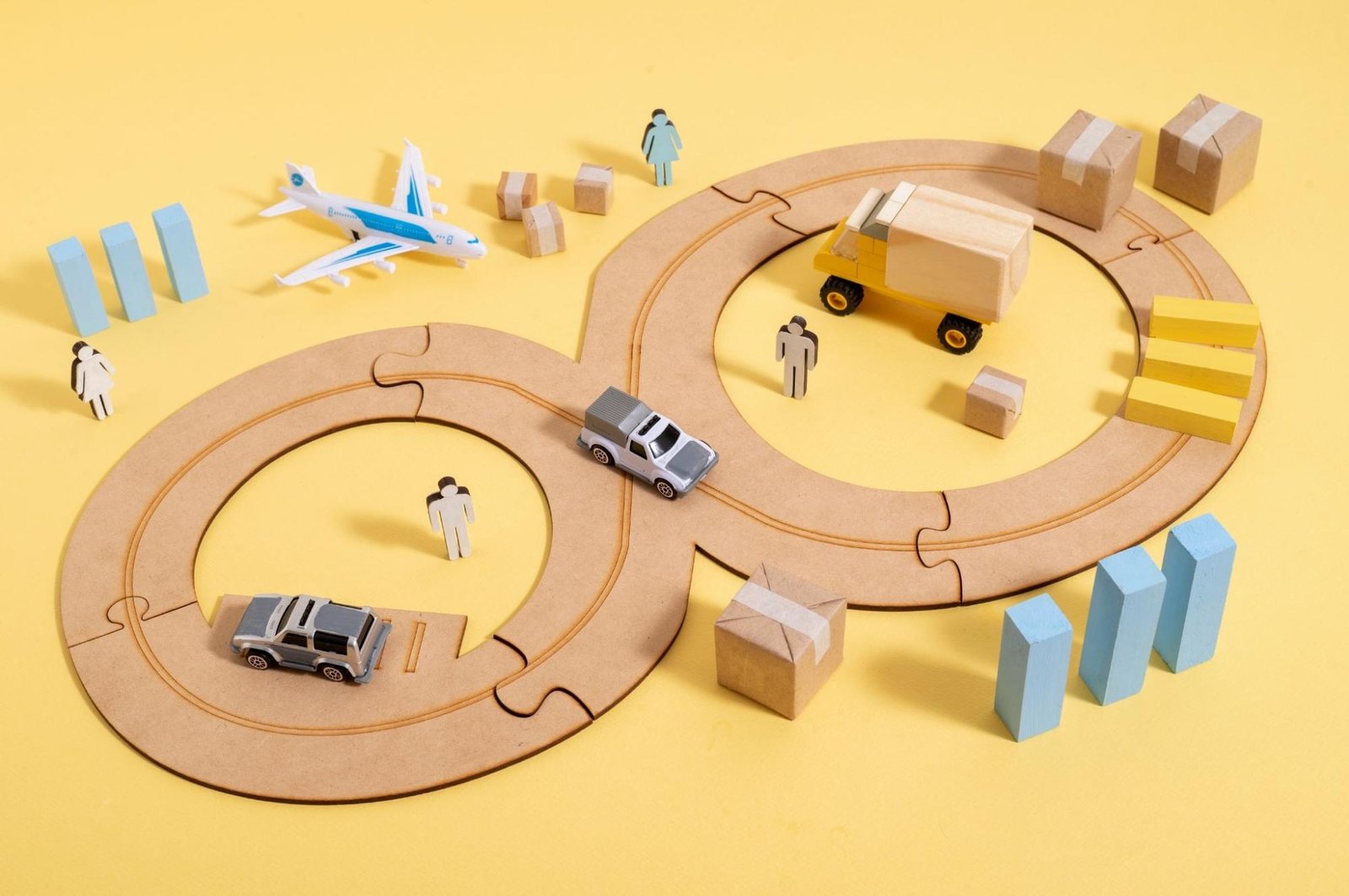 Toy model representing logistics and transportation on a yellow background. The setup includes a wooden infinity-shaped road track with miniature vehicles (truck and cars), cardboard boxes symbolizing parcels, small human figures, a toy airplane, and block buildings. The scene illustrates the flow of goods and supply chain concepts.