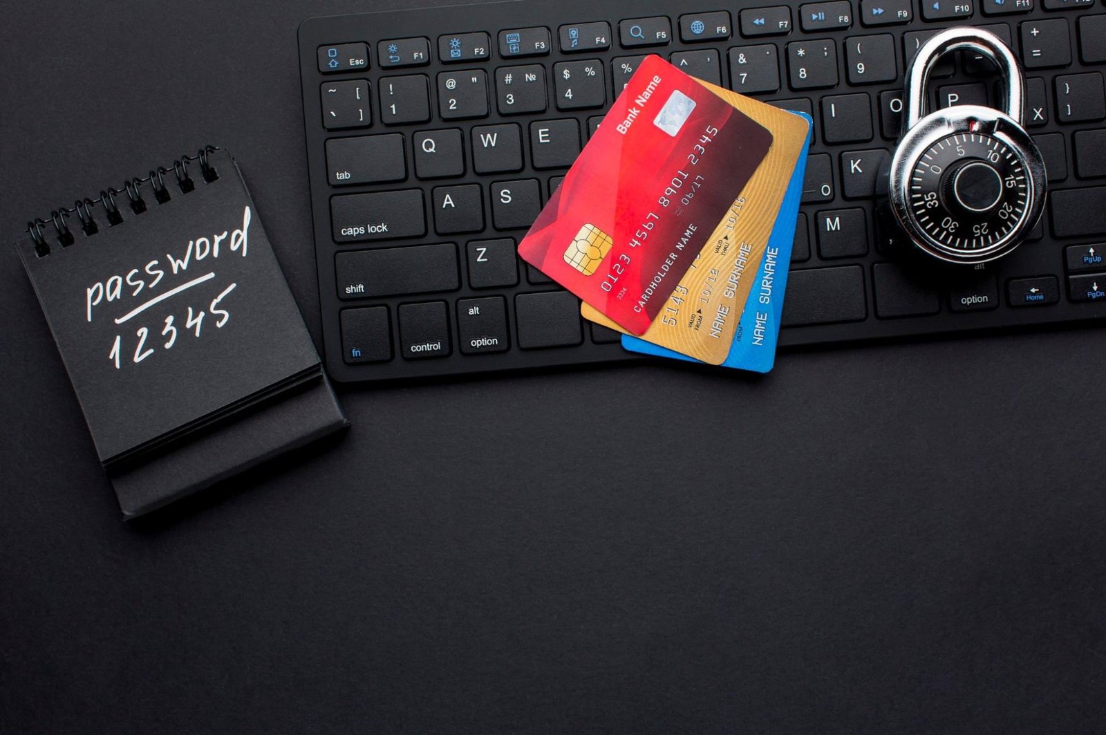 A flat lay shows a keyboard, a notebook with "password 12345", stacked red and blue credit cards, and an open padlock on a black background, illustrating cybersecurity and password safety.